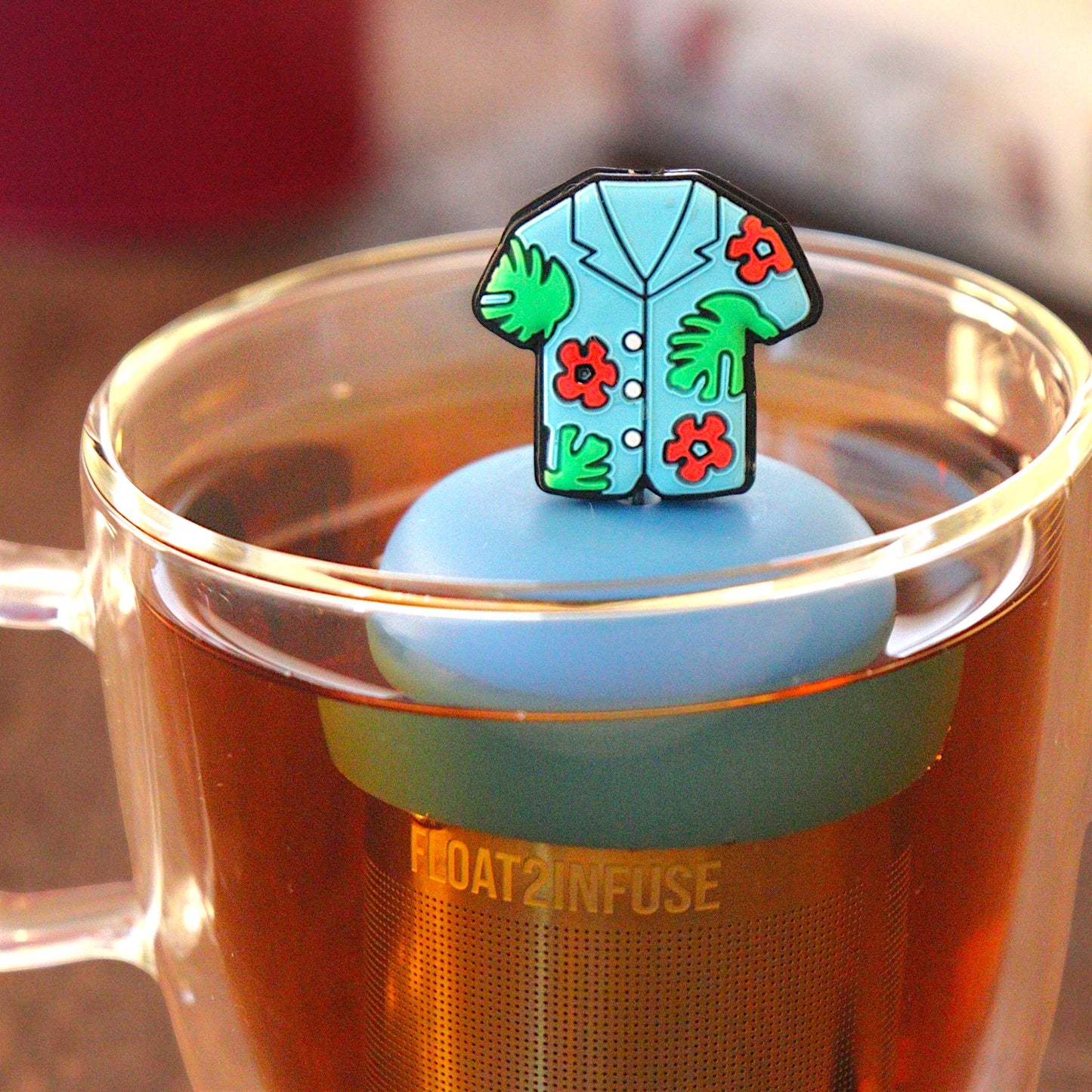 blue Hawaiian shirt tea infuser for loose leaf tea  with a fine mesh stainless steel tea strainer that reads FLOAT2INFUSE on a blue base floating in a glass tea mug with a loose leaf tea and books in the background