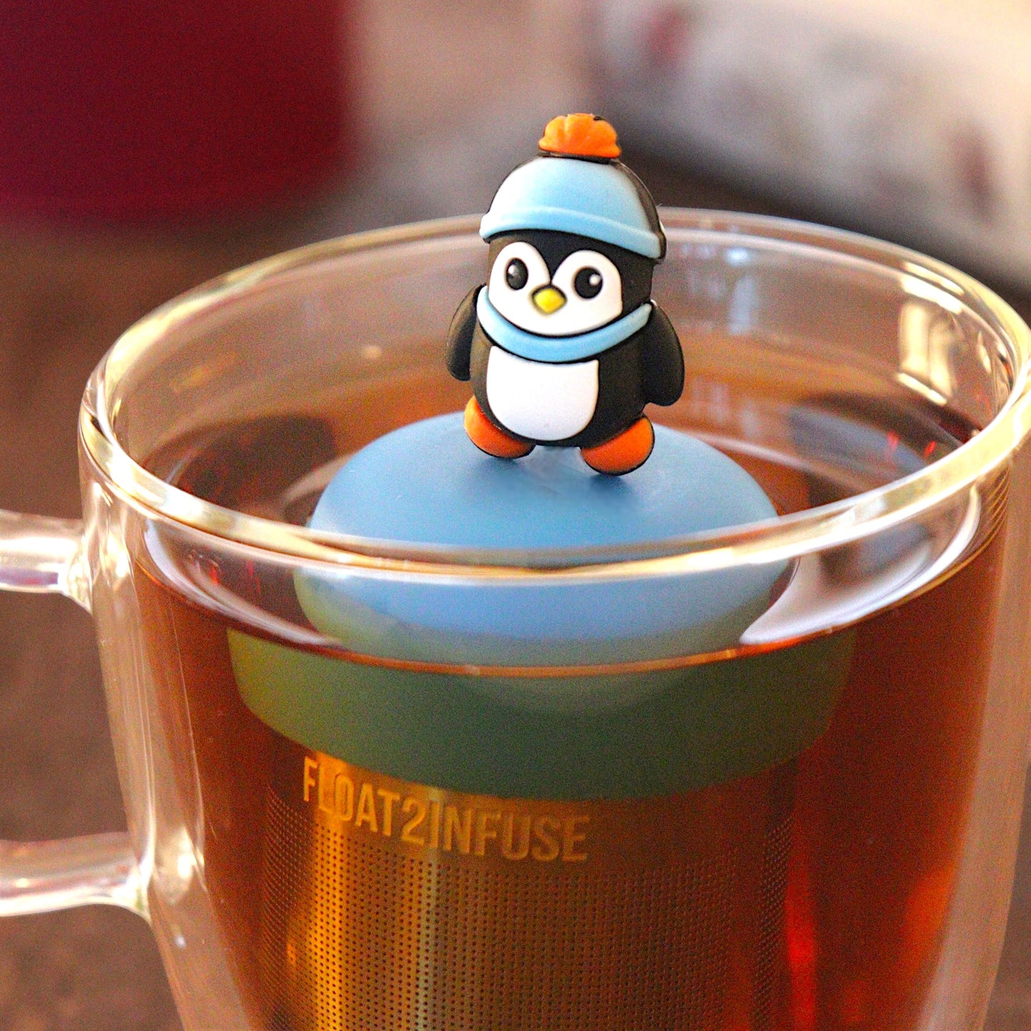 cute animal penguin wearing a blue hat and scarf tea infuser for loose leaf tea with a fine mesh stainless steel tea infuser that reads FLOAT2INFUSE on a blue base  floating in a glass tea mug with  loose leaf tea andbooks in the background
