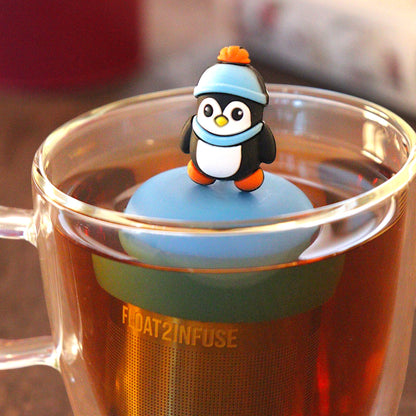 cute animal penguin wearing a blue hat and scarf tea infuser for loose leaf tea with a fine mesh stainless steel tea infuser that reads FLOAT2INFUSE on a blue base  floating in a glass tea mug with  loose leaf tea andbooks in the background