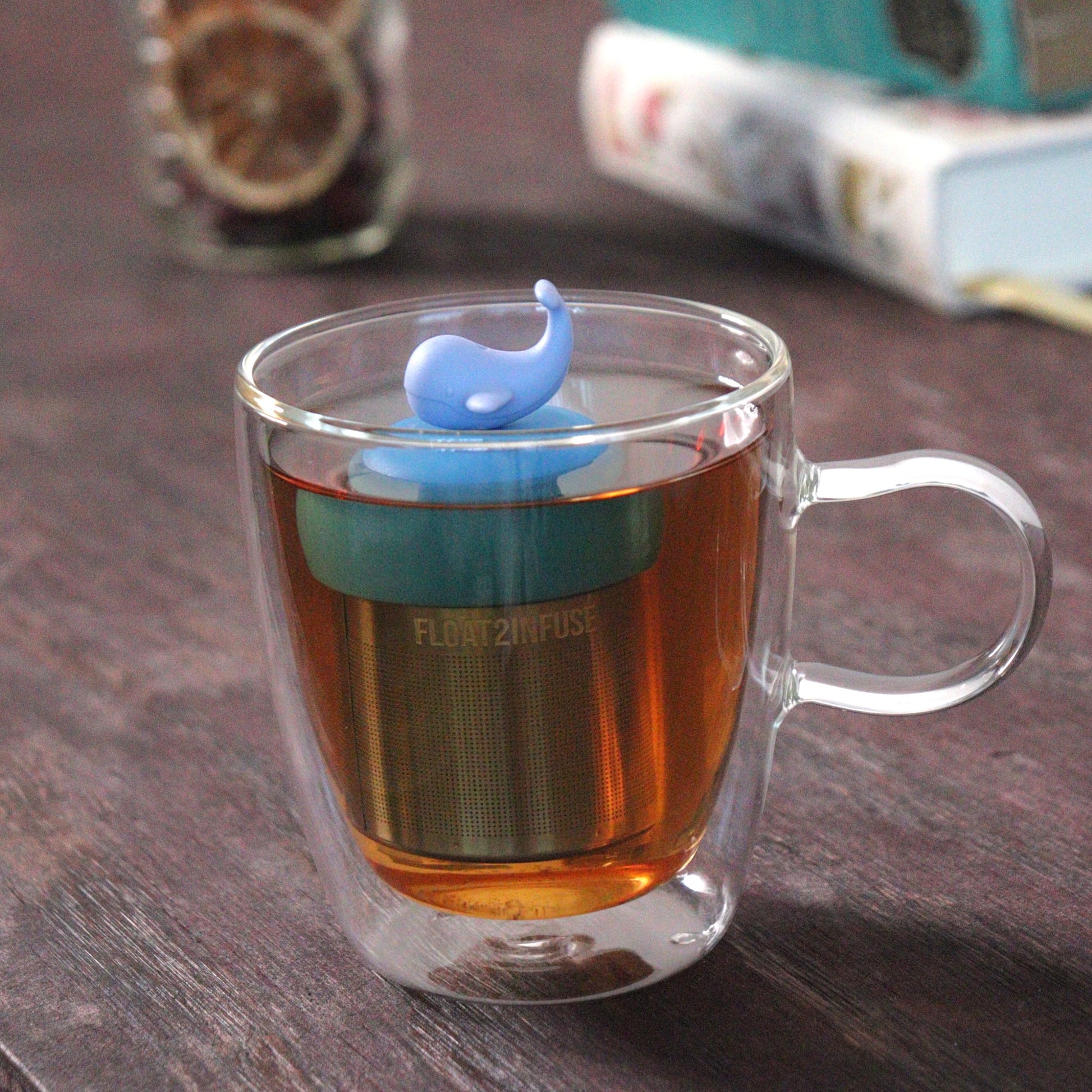 whale tea infuser for loose leaf tea  with a fine mesh stainless steel tea strainer that reads FLOAT2INFUSE on a blue base floating in a glass tea mug with loose leaf tea and books in the background