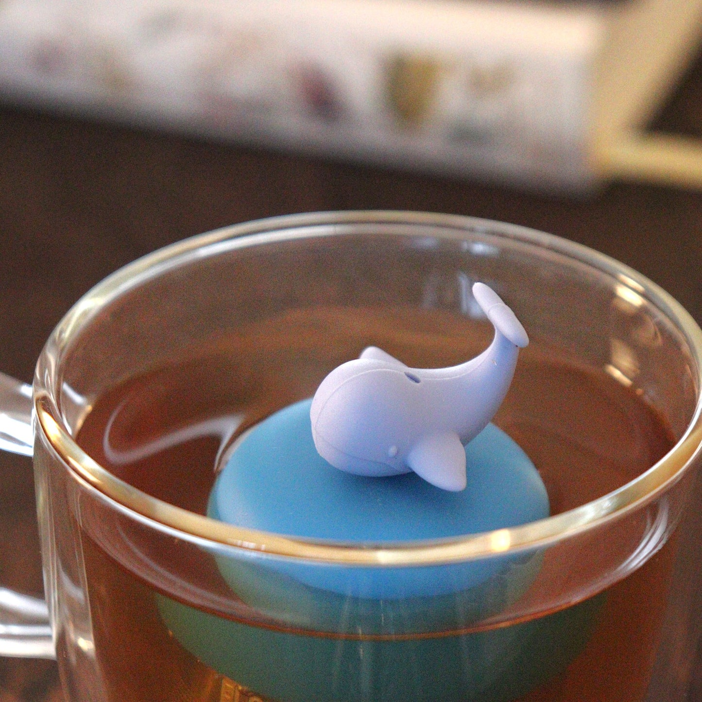 whale tea infuser for loose leaf tea on a blue base with a fine mesh stainless steel tea strainer that reads FLOAT2INFUSE floating in a glass tea mug with books in the background