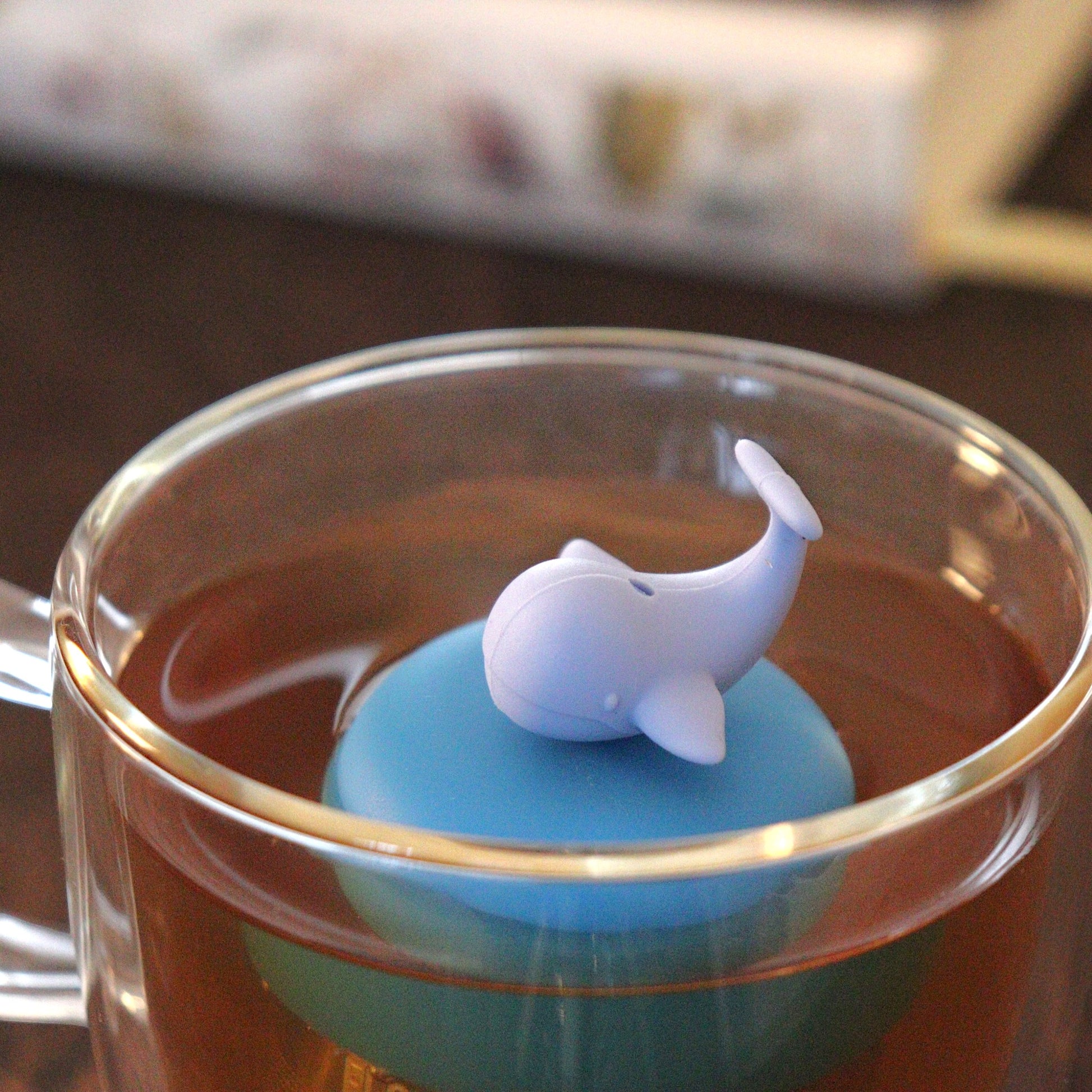 whale tea infuser for loose leaf tea on a blue base with a fine mesh stainless steel tea strainer that reads FLOAT2INFUSE floating in a glass tea mug with books in the background