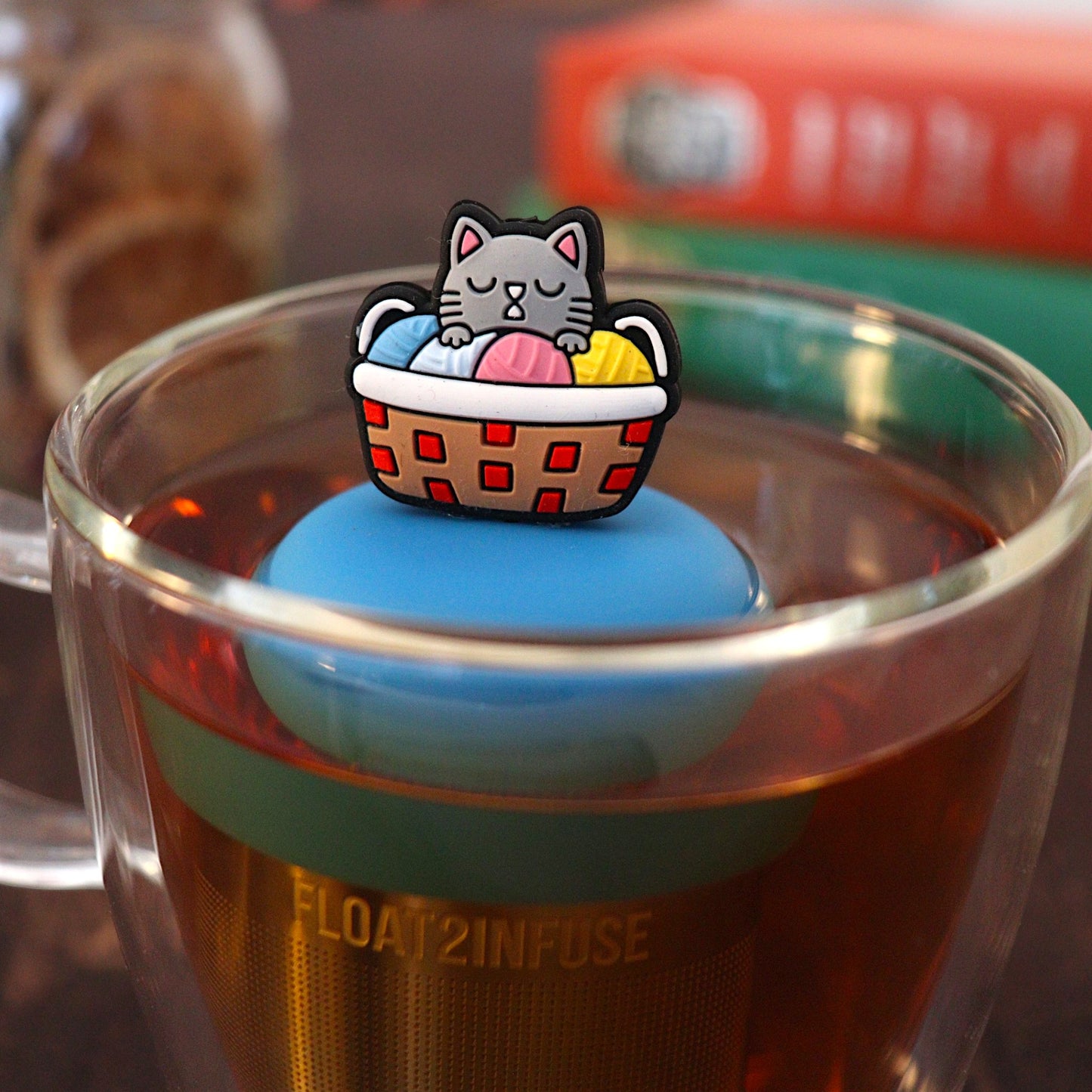 grey cat in colorful knitting basket animal  tea infuser for Loose leaf tea with a fine mesh stainless steel Tea steeper that reads FLOAT2INFUSE on blue base floating in a Glass tea mug with books and loost leaf tea in the background