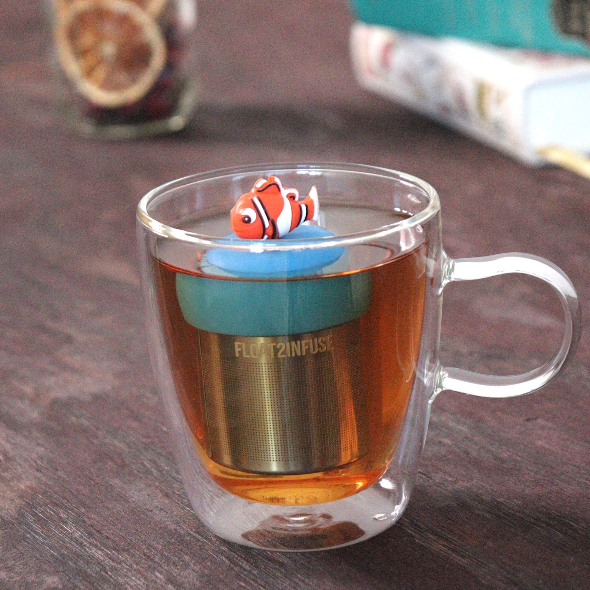 clownfish animal tea infuser for loose leaf tea with a fine mesh stainless steel tea strainer that reads FLOAT2INFUSE  on a blue base  floating in a glass tea mug with loose leaf tea and books in the background