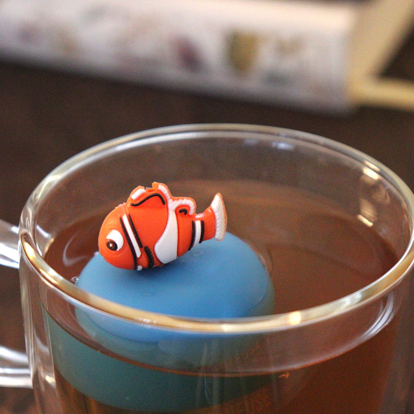 clownfish animal tea infuser for loose leaf tea  with a fine mesh stainless steel tea strainer that reads FLOAT2INFUSE on a blue base floating in a glass tea mug with books in the background