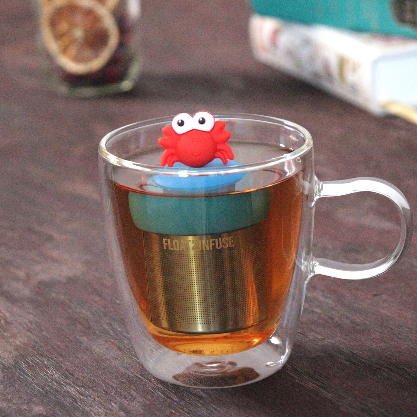red crab animal tea infuser for loose leaf tea  with a fine mesh stainless steel tea strainer that reads FLOAT2INFUSE on a blue base floating in a glass tea mug with loose leaf tea and books in the background