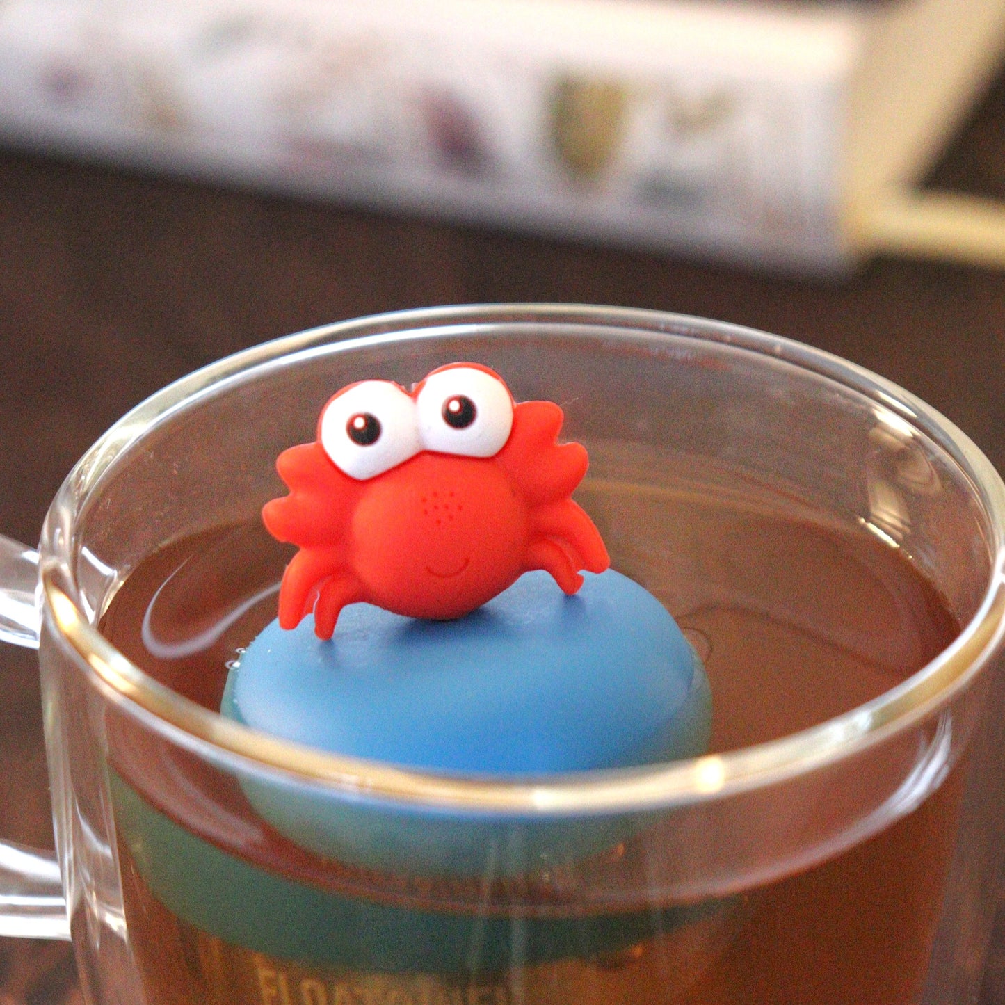 red crab animal tea infuser for loose leaf tea with a fine mesh stainless steel tea strainer that reads FLOAT2INFUSE  on a blue base floating in a glass tea mug with books in the background