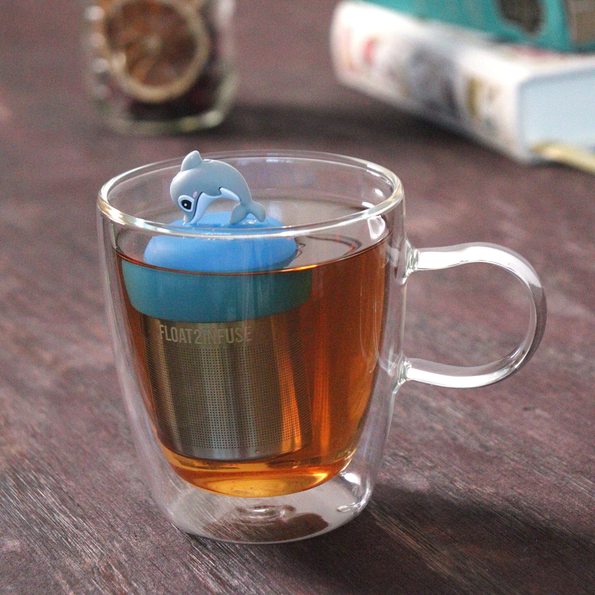 cute dolphin animal tea infuser for loose leaf tea with a fine mesh stainless steel tea strainer that reads FLOAT2INFUSE  on a blue base floating in a glass tea mug with loose leaf tea and books in the background