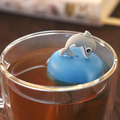 cute dolphin animal tea infuser for loose leaf tea  with a fine mesh stainless steel tea strainer that reads FLOAT2INFUSE on a blue base floating in a glass tea mug with books in the background
