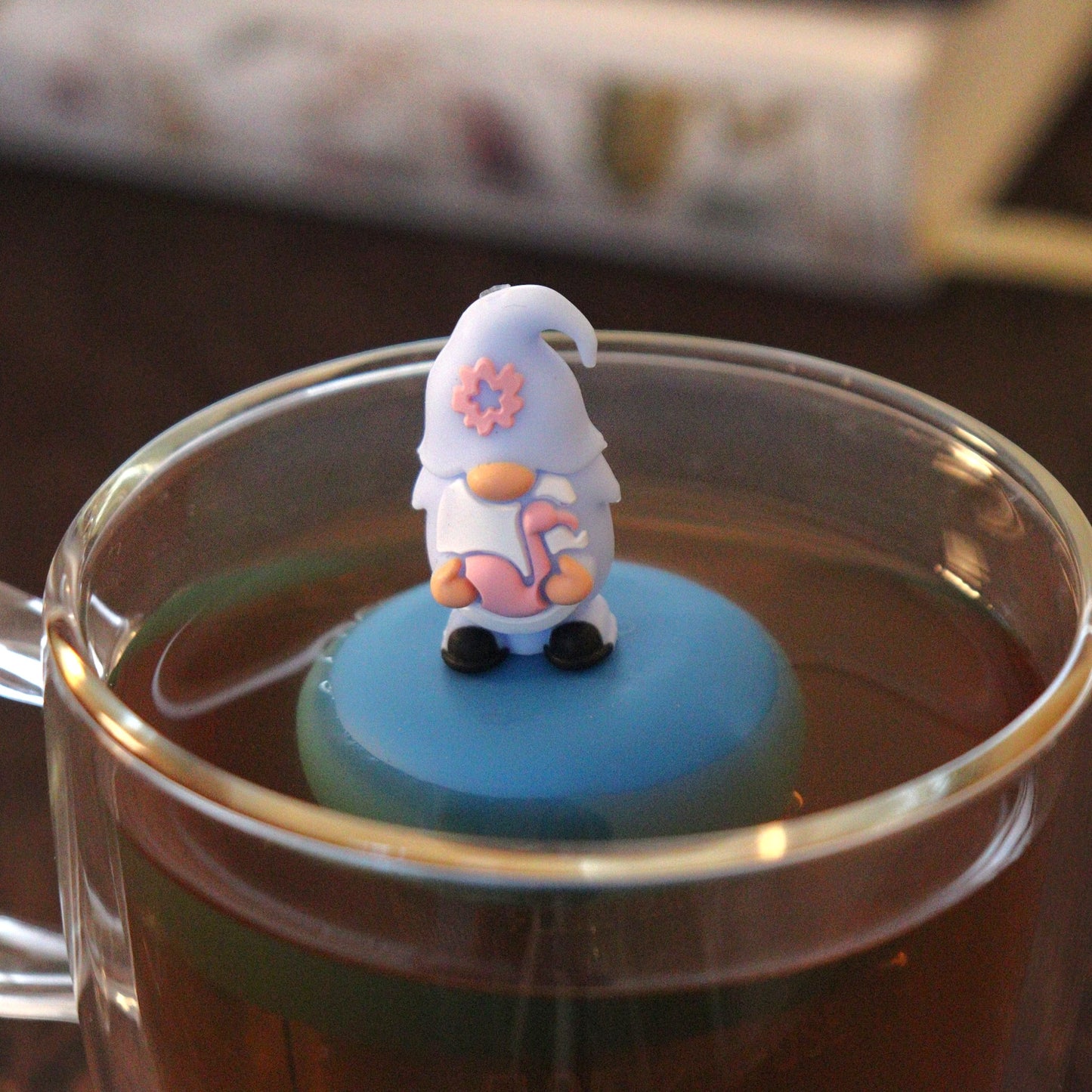 lavender gnome holding flamingo animal tea infuser for loose leaf tea  with a fine mesh stainless steel tea strainer that reads FLOAT2INFUSE on a blue base floating in a glass tea mug with books in the background