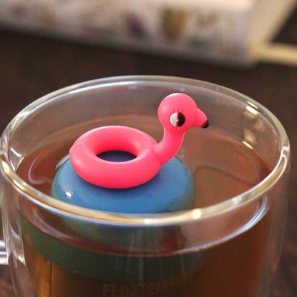 cute pink flamingo pool float tea infuser for loose leaf tea with a fine mesh stainless steel tea strainer that reads FLOAT2INFUSE on a blue base floating in a glass tea mug with books in the background