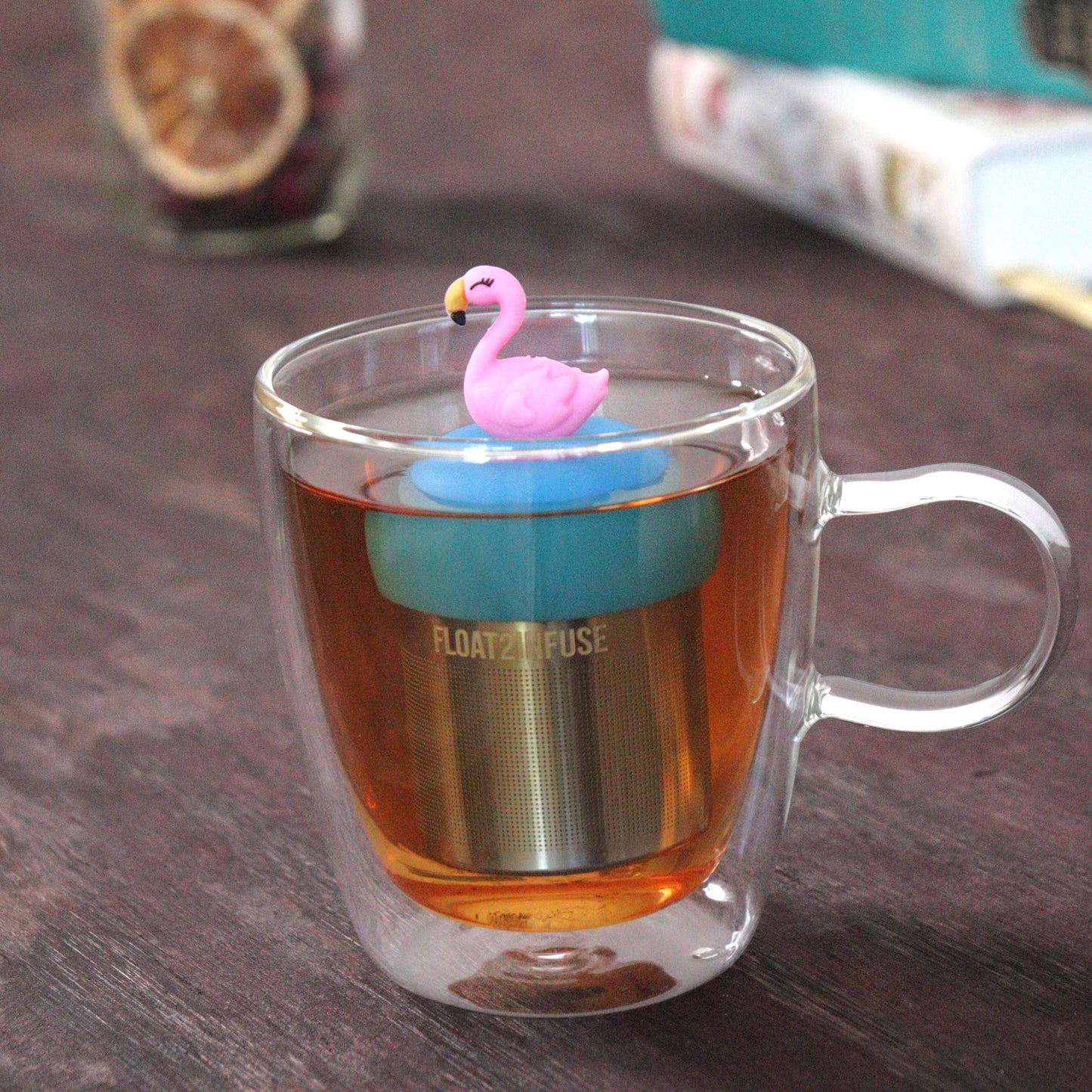 pink flamingo tea infuser for loose leaf tea with a fine mesh stainless steel tea strainer that reads FLOAT2INFUSE on a blue base floating in a glass tea mug with loose leaf tea and books in the background