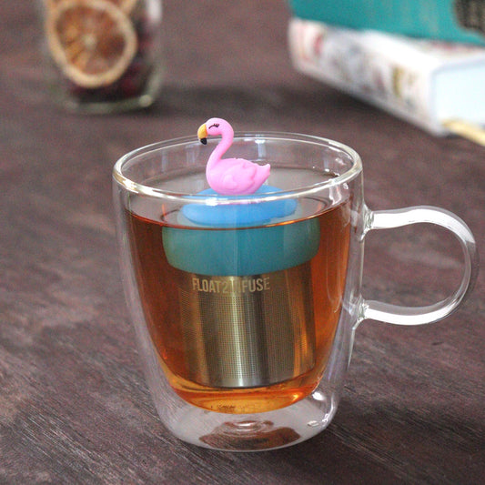 pink flamingo tea infuser for loose leaf tea with a fine mesh stainless steel tea strainer that reads FLOAT2INFUSE on a blue base floating in a glass tea mug with loose leaf tea and books in the background