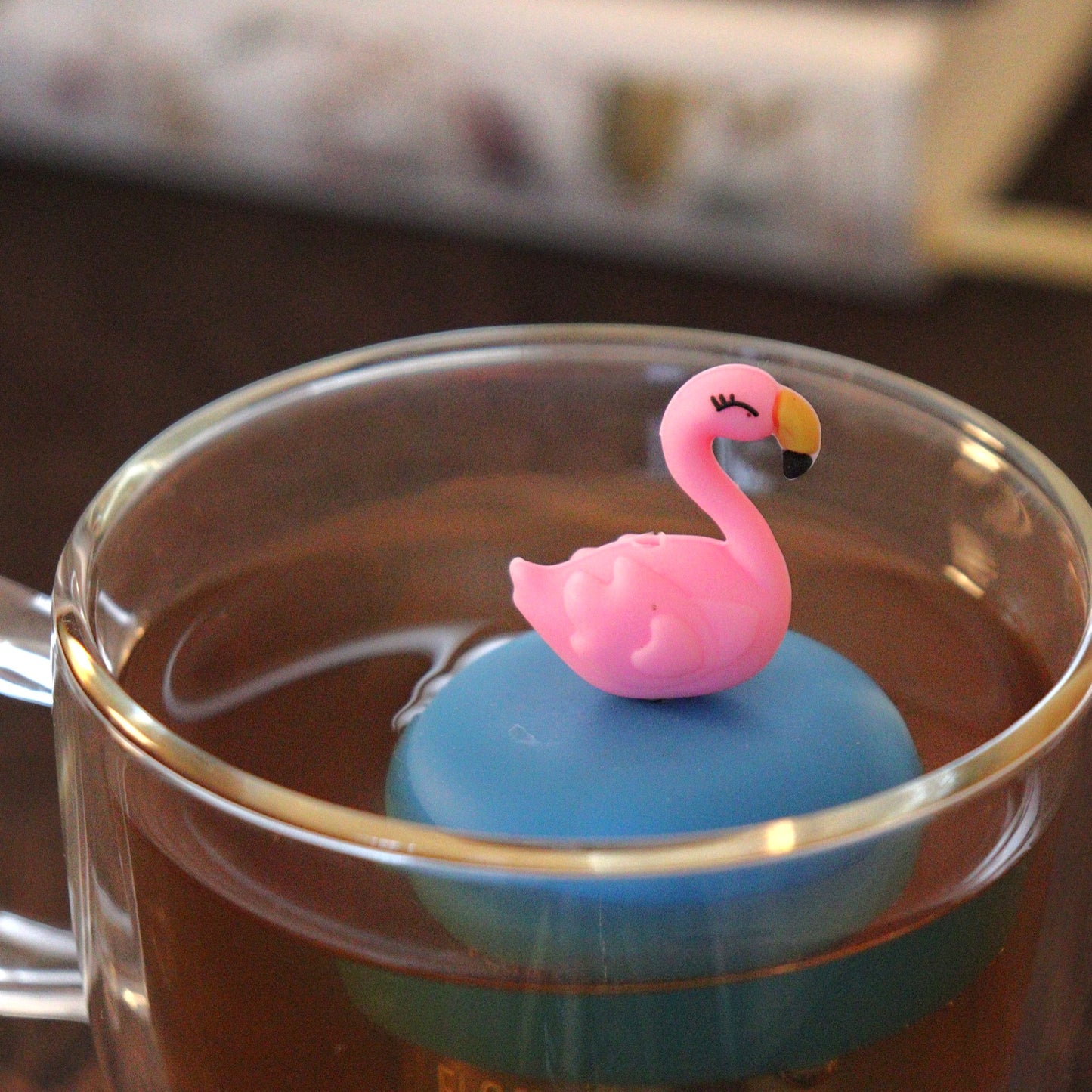 pink flamingo tea infuser for loose leaf tea with a fine mesh stainless steel tea strainer that reads FLOAT2INFUSE on a blue base floating in a glass tea mug with books in the background