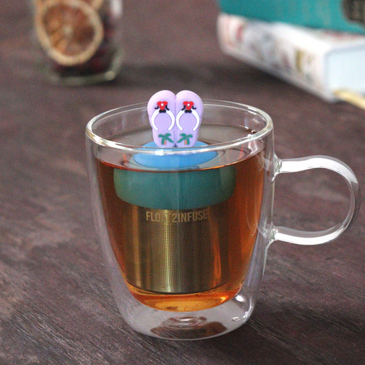 lavender beach flip flop tea infuser for loose leaf tea on a blue base with a fine mesh stainless steel tea strainer that reads FLOAT2INFUSE floating in a glass tea mug with loose leaf tea and books in the background