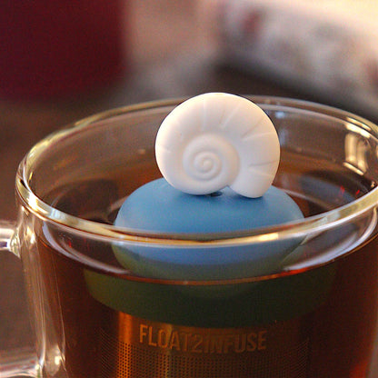 Fossil sea shell tea infuser for loose leaf tea  with a fine mesh stainless steel tea strainer that reads FLOAT2INFUSE on a blue base floating in a glass tea mug with loose leaf tea and books in the background