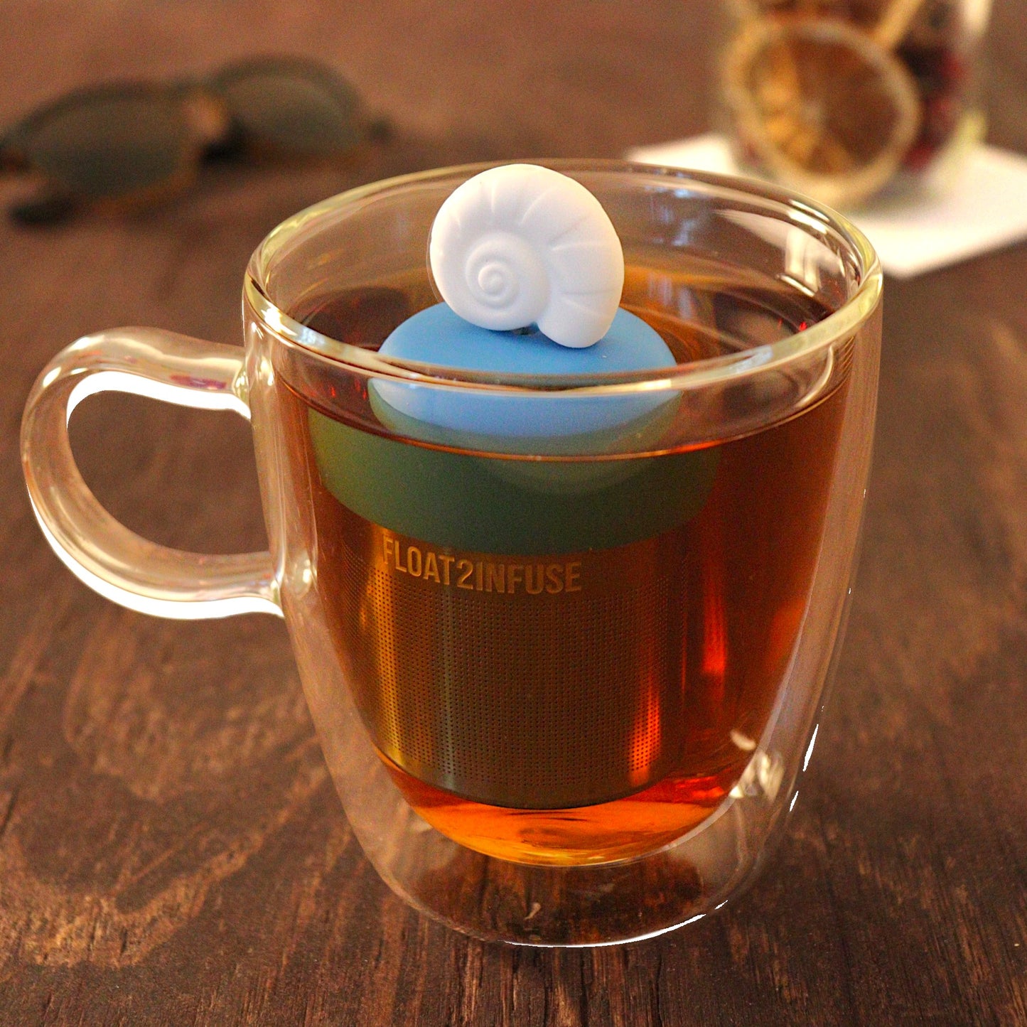 Fossil sea shell tea infuser for loose leaf tea  with a fine mesh stainless steel tea strainer that reads FLOAT2INFUSE on a blue base floating in a glass tea mug with loose leaf tea and sunglasses in the background