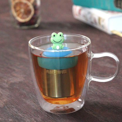 cute green animal tea infuser for loose leaf tea  with a fine mesh stainless steel tea strainer that reads FLOAT2INFUSE on a blue base floating in a glass tea mug with loose leaf tea and books in the background