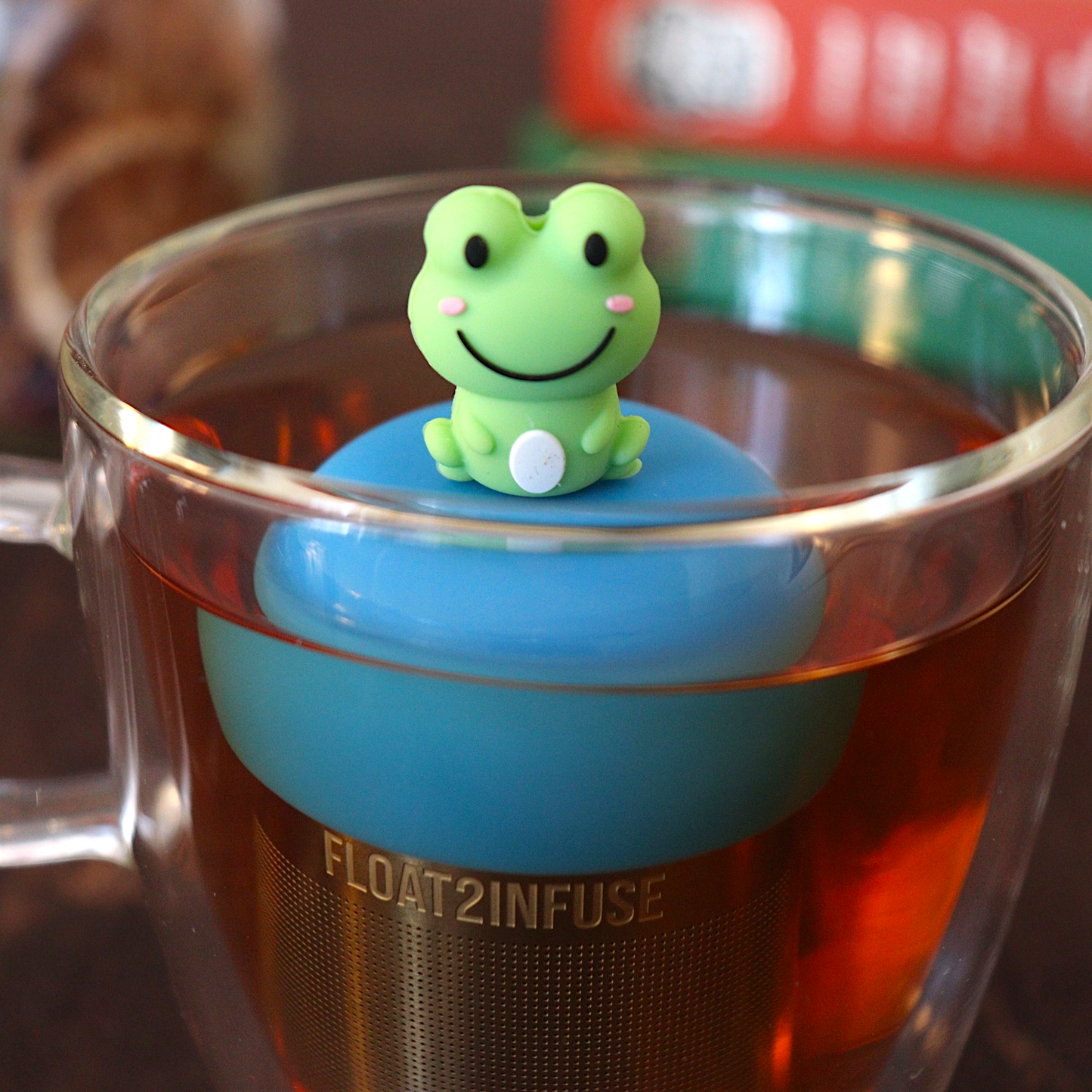 cute green frog animal tea infuser for loose leaf tea with a fine mesh stainless steel tea strainer that reads FLOAT2INFUSE on a blue base floating in a glass tea mug with books in the background