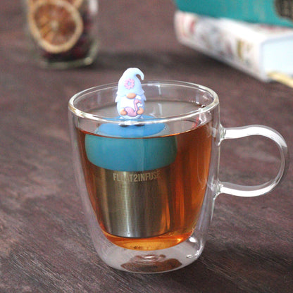 lavender gnome holding flamingo animal tea infuser for loose leaf tea  with a fine mesh stainless steel tea strainer that reads FLOAT2INFUSE on a blue base floating in a glass tea mug with loose leaf tea and books in the background