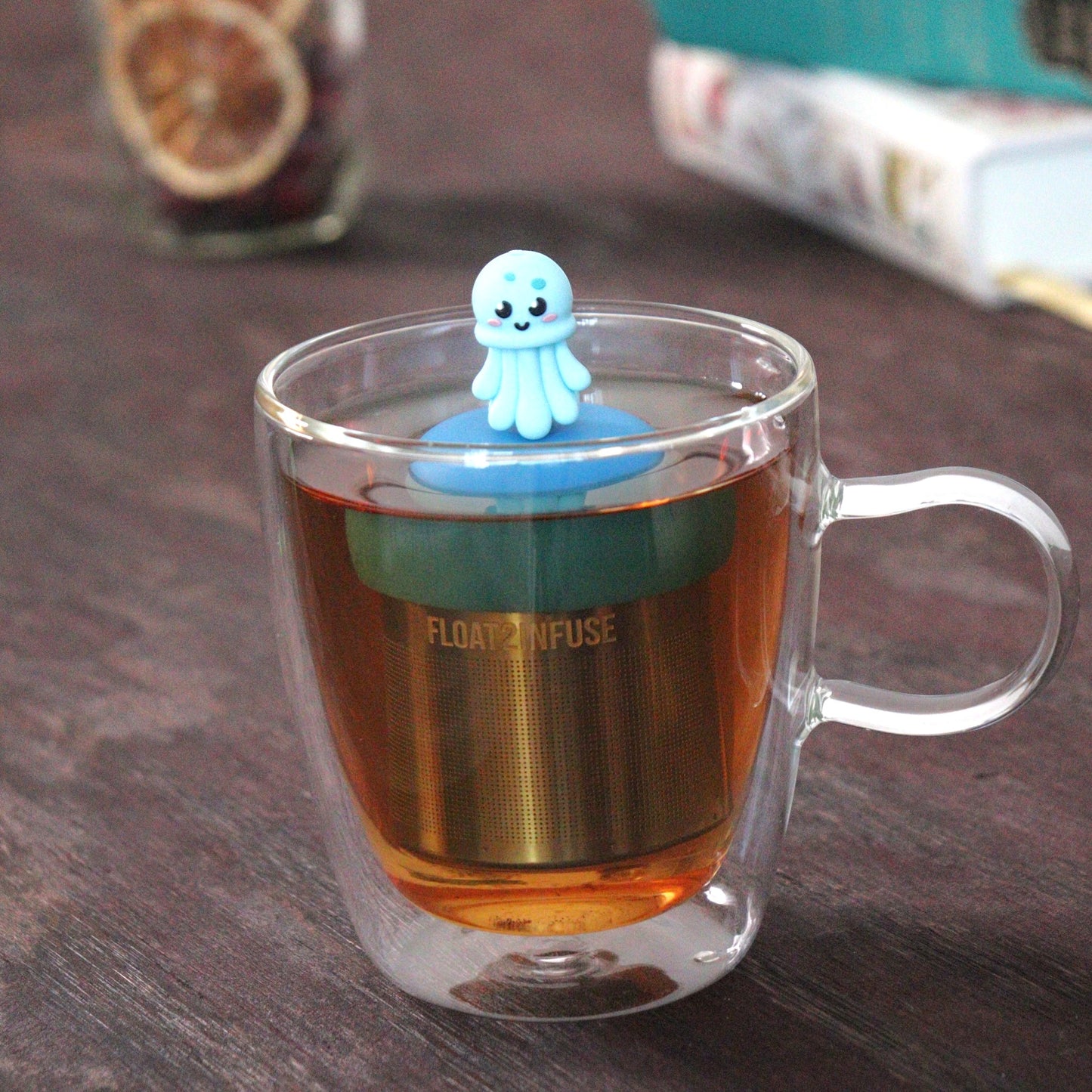 blue jellyfish animal tea infuser for loose leaf tea with a fine mesh stainless steel tea strainer that reads FLOAT2INFUSE on a blue base  floating in a glass tea mug with loose leaf tea and books in the background