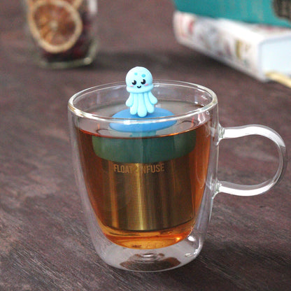 blue jellyfish animal tea infuser for loose leaf tea with a fine mesh stainless steel tea strainer that reads FLOAT2INFUSE on a blue base  floating in a glass tea mug with loose leaf tea and books in the background