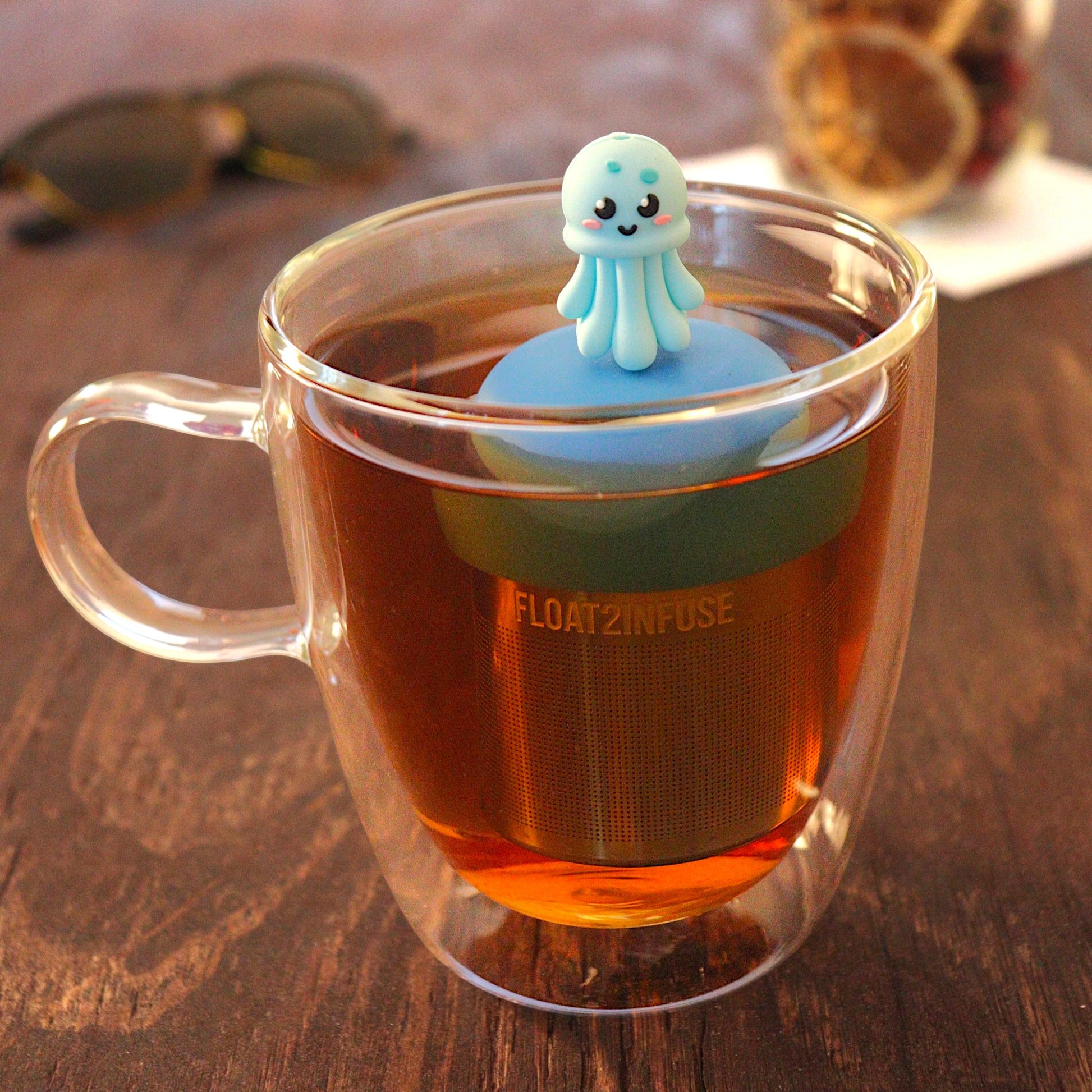 blue jellyfish animal tea infuser for loose leaf tea  with a fine mesh stainless steel tea strainer that reads FLOAT2INFUSE on a blue base floating in a glass tea mug with loose leaf tea and sunglasses in the background