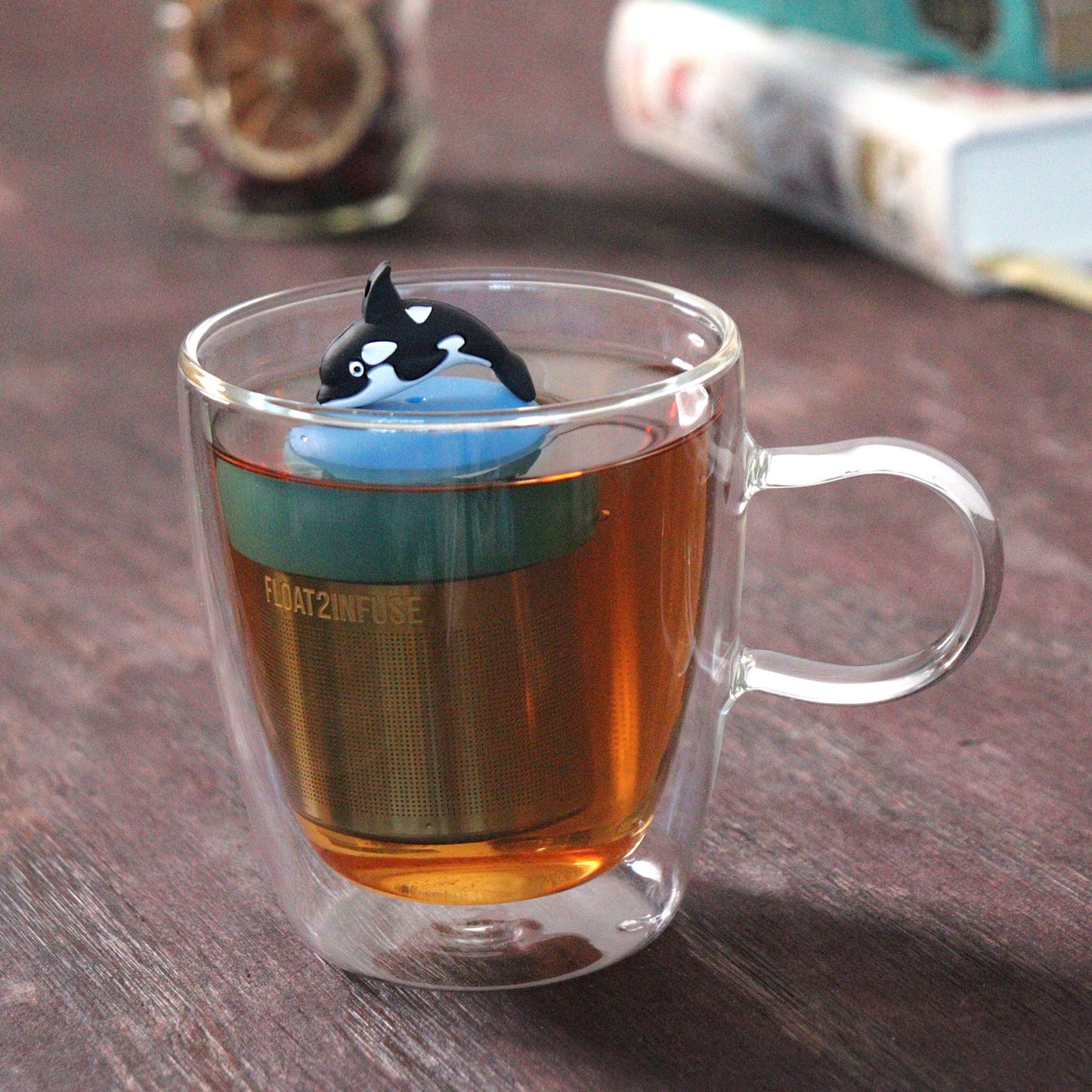 killer whale animal tea infuser for loose leaf tea  with a fine mesh stainless steel tea strainer that reads FLOAT2INFUSE on a blue base floating in a glass tea mug with loose leaf tea and books in the background