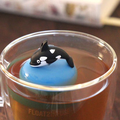 killer whale animal tea infuser for loose leaf tea  with a fine mesh stainless steel tea strainer that reads FLOAT2INFUSE on a blue base floating in a glass tea mug with books in the background