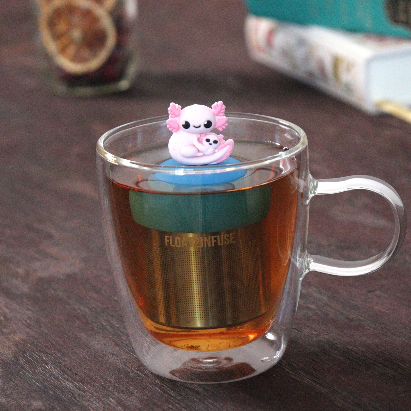 pink axolotl mom and baby tea infuser for loose leaf tea with a fine mesh stainless steel tea strainer that reads FLOAT2INFUSE on a blue base  floating in a glass tea mug with loose leaf tea and books in the background