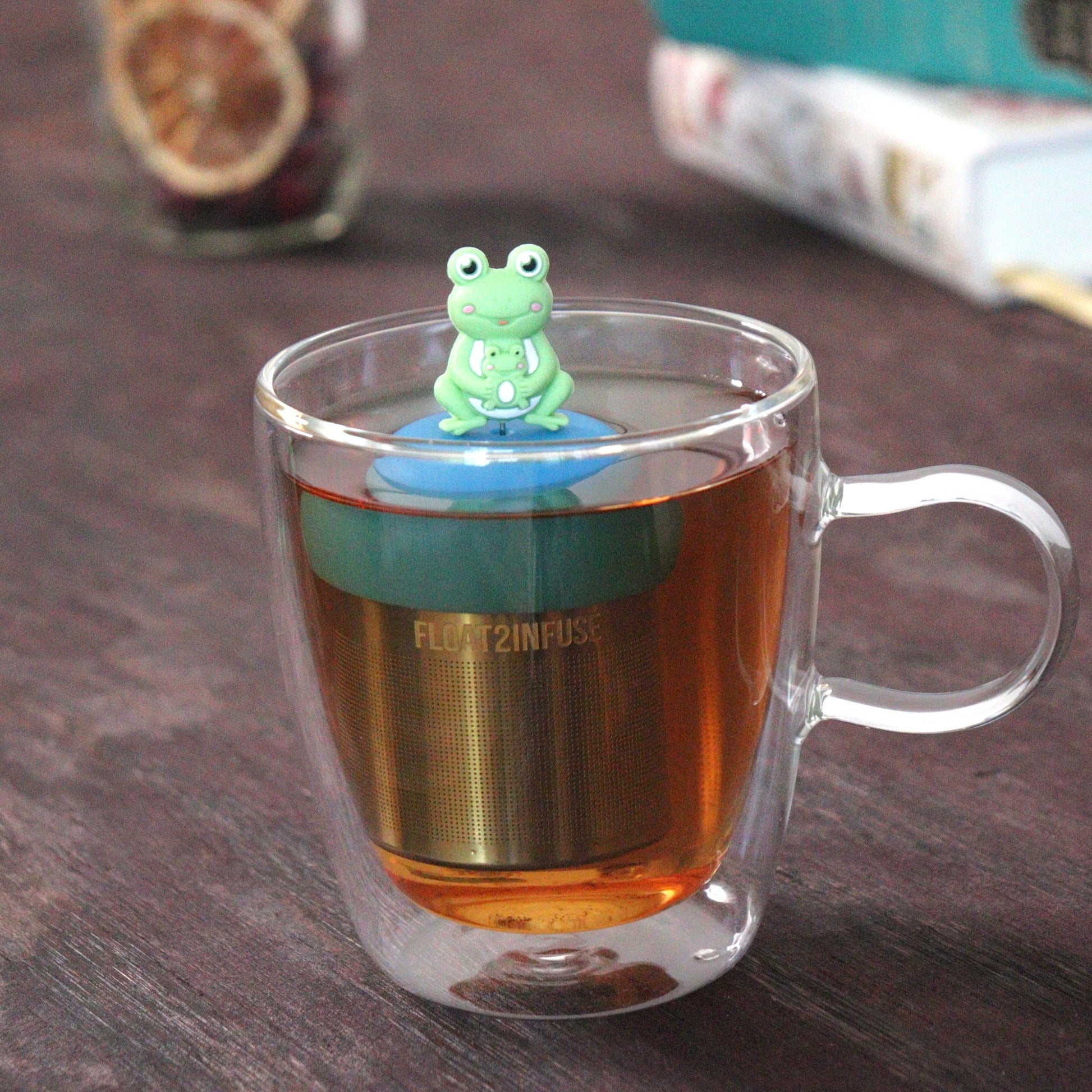 green frog mom and baby animal tea infuser for loose leaf tea  with a fine mesh stainless steel tea strainer that reads FLOAT2INFUSE on a blue base floating in a glass tea mug with loose leaf tea and books in the background