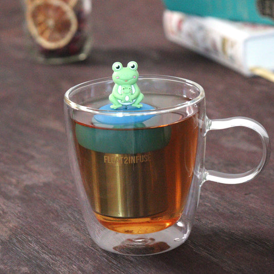 green frog mom and baby animal tea infuser for loose leaf tea on a blue base with a fine mesh stainless steel tea strainer that reads FLOAT2INFUSE floating in a glass tea mug with loose leaf tea and books in the background