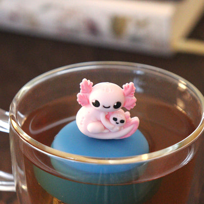 pink axolotl mom and baby animal tea infuser for loose leaf tea with a fine mesh stainless steel tea strainer that reads FLOAT2INFUSE on a blue base  floating in a glass tea mug with books in the background