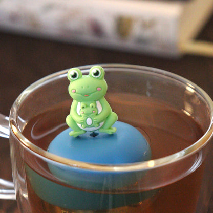 green frog mom and baby animal tea infuser for loose leaf tea  with a fine mesh stainless steel tea strainer that reads FLOAT2INFUSE on a blue base floating in a glass tea mug with books in the background