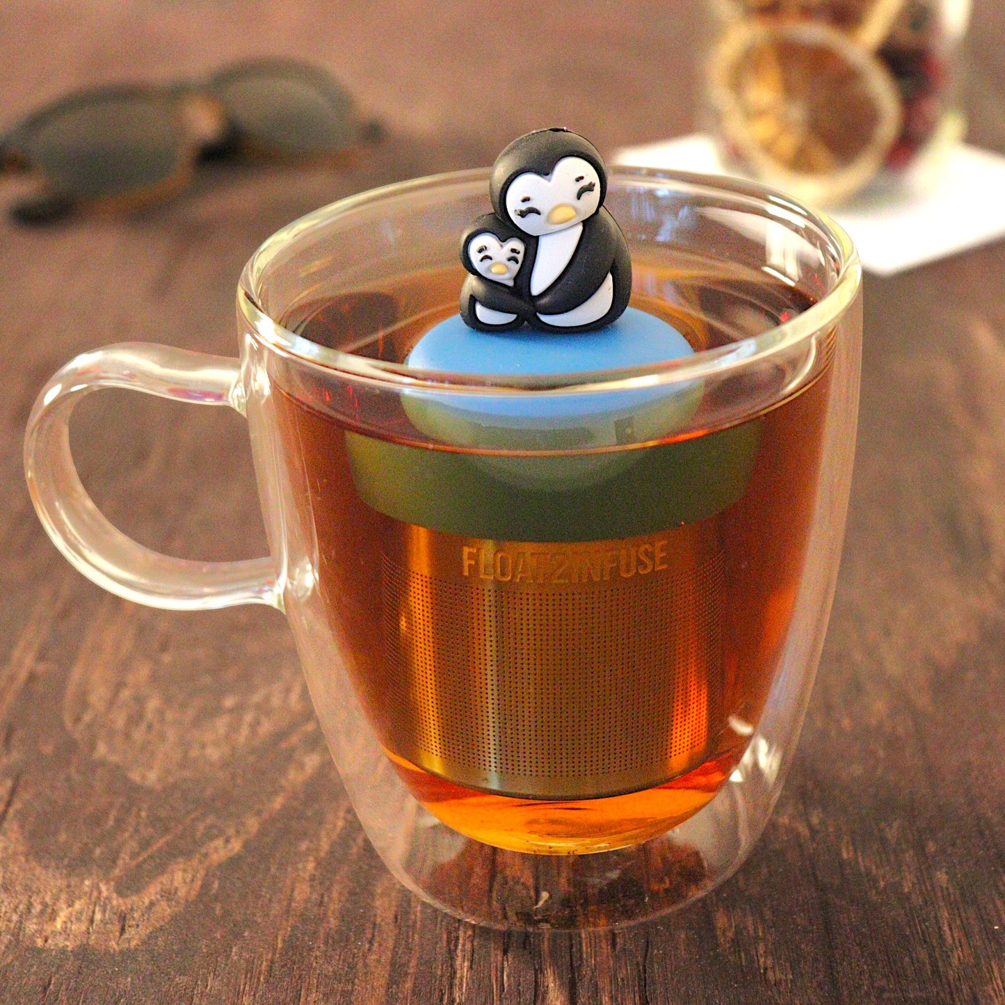 Black and white mom and baby penguin hugging tea infuser for loose leaf tea with a fine mesh stainless steel tea infuser that reads FLOAT2INFUSE on a blue base floating in a glass tea mug with a sunglasses in the background