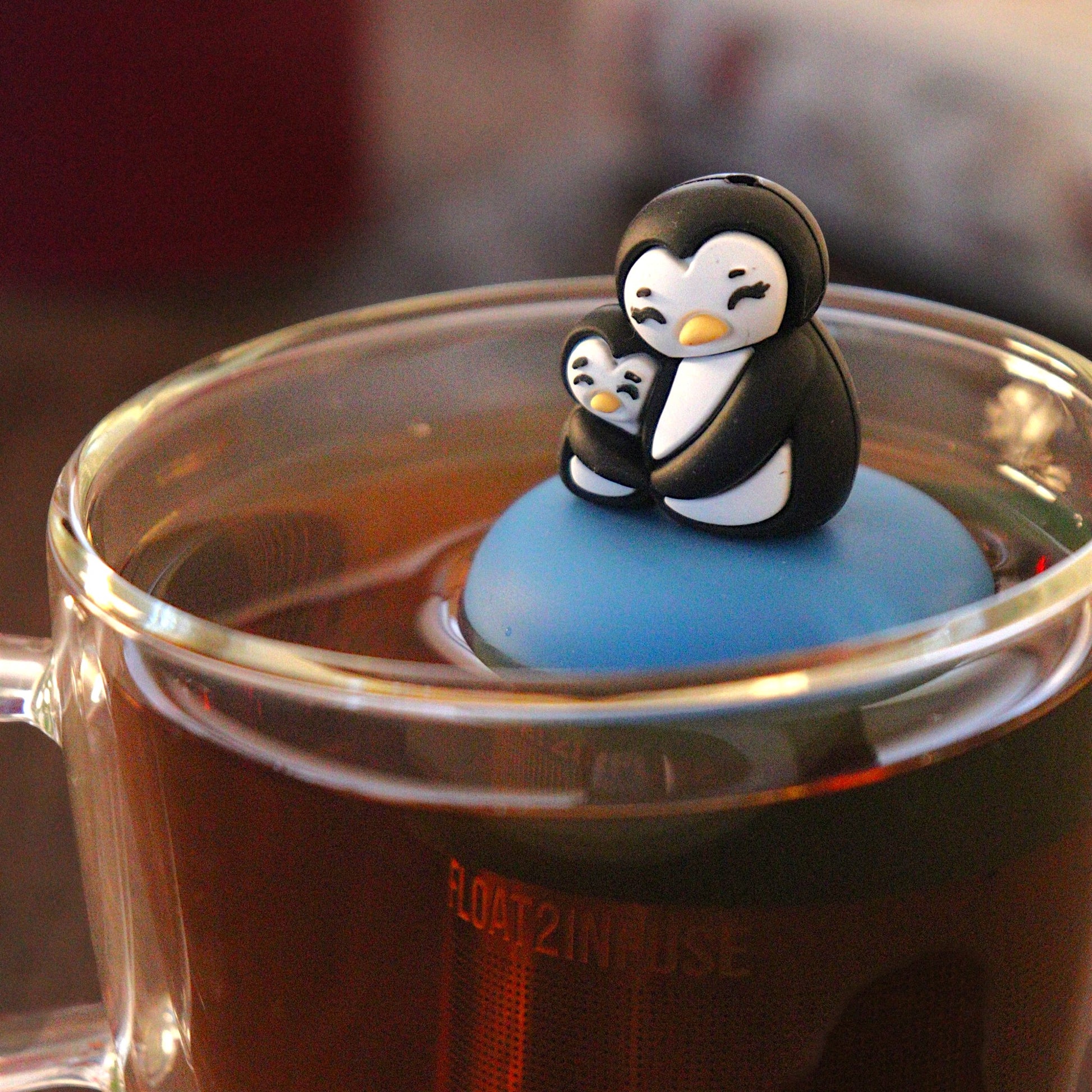 Black and white mom and baby penguin hugging tea infuser for loose leaf tea with a fine mesh stainless steel tea infuser that reads FLOAT2INFUSE on a blue base floating in a glass tea mug 