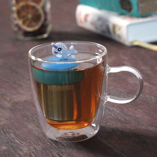 cute grey narwal animal tea infuser for loose leaf tea with a fine mesh stainless steel tea strainer that reads FLOAT2INFUSE on a blue base  floating in a glass tea mug with loose leaf tea and books in the background