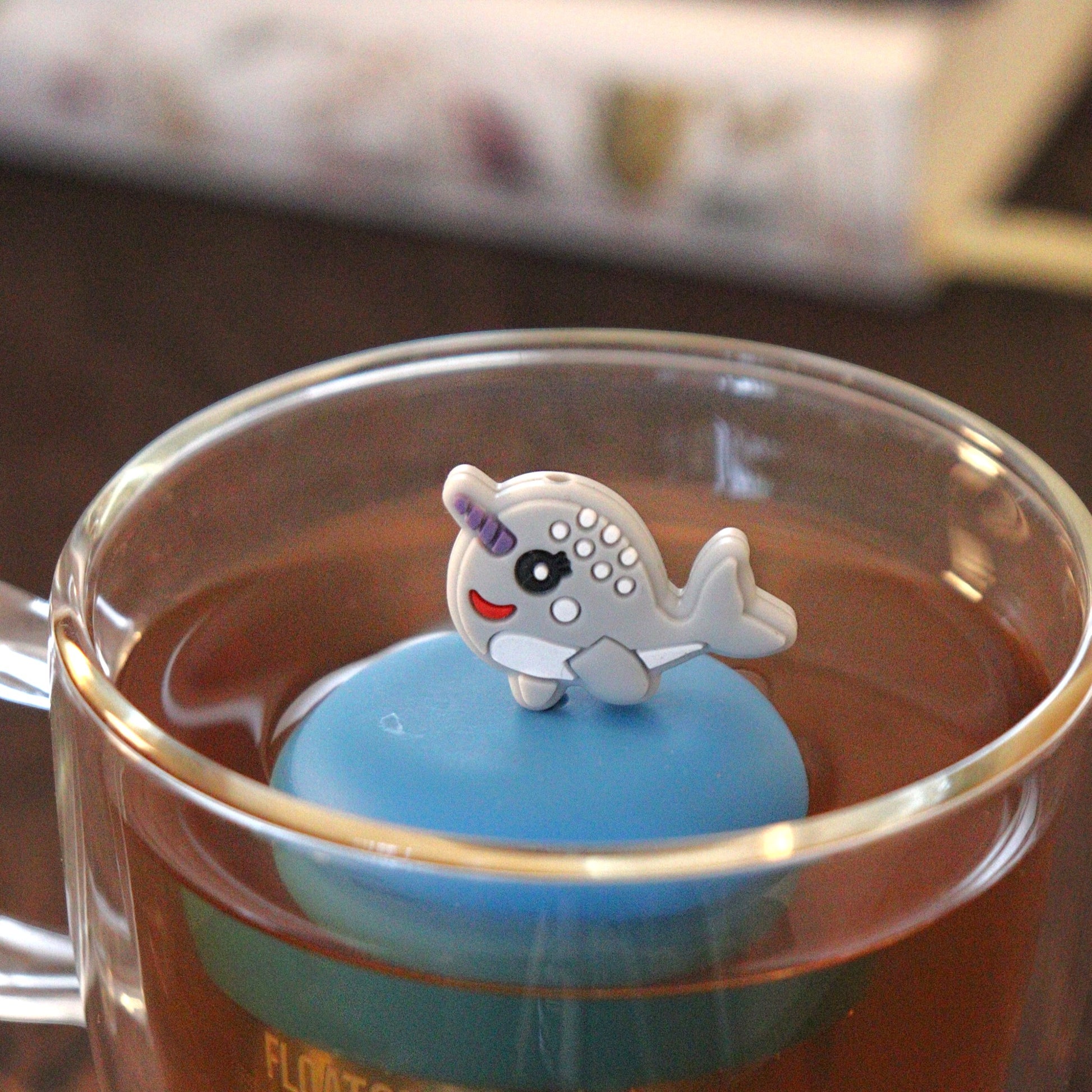 cute grey narwhal animal tea infuser for loose leaf tea on a blue base with a fine mesh stainless steel tea strainer that reads FLOAT2INFUSE floating in a glass tea mug with books in the background