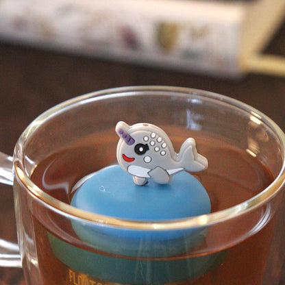 cute grey narwhal animal tea infuser for loose leaf tea on a blue base with a fine mesh stainless steel tea strainer that reads FLOAT2INFUSE floating in a glass tea mug with books in the background