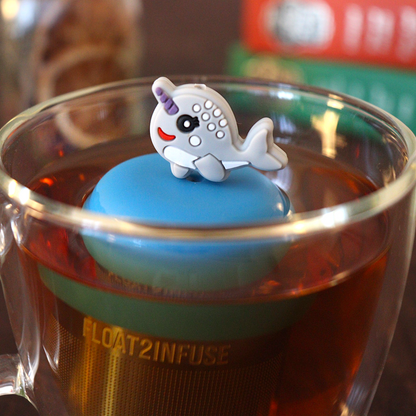 cute grey narwhal animal tea infuser for loose leaf tea on a blue base with a fine mesh stainless steel tea strainer that reads FLOAT2INFUSE floating in a glass tea mug with books in the background