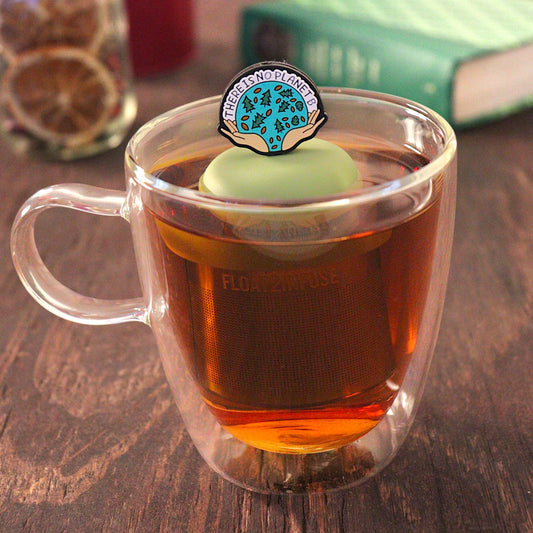 black circle with open hands holding images of leaves and pine trees, and text that reads "There is no planet b"  tea infuser for loose leaf tea on a green base with a fine mesh stainless steel tea infuser that reads FLOAT2INFUSE floating in a glass tea mug with pen on notebook and loose leaf tea and books in the background