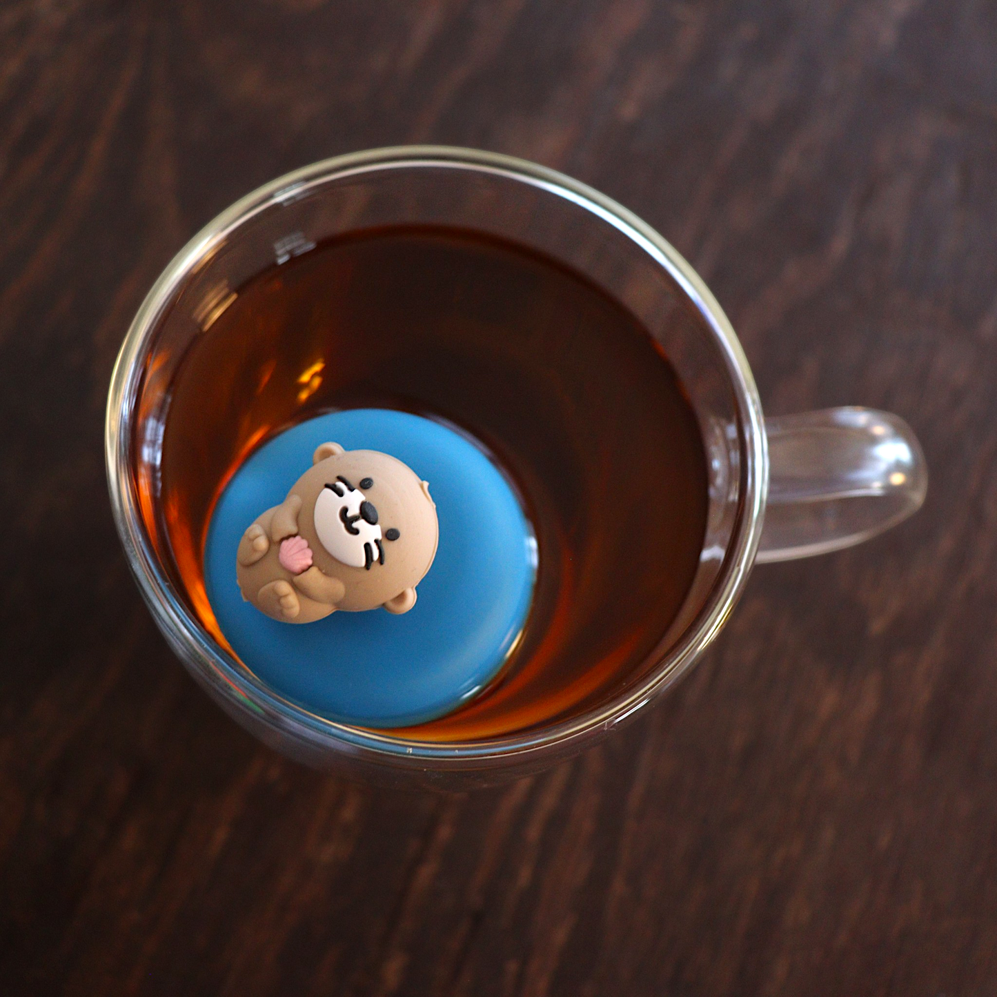Looking down at a cute little otter floating on it's back holding a pink seashell tea infuser for loose leaf tea with a fine mesh stainless steel tea strainer that reads FLOAT2INFUSE on a blue base floating in a glass tea mug 