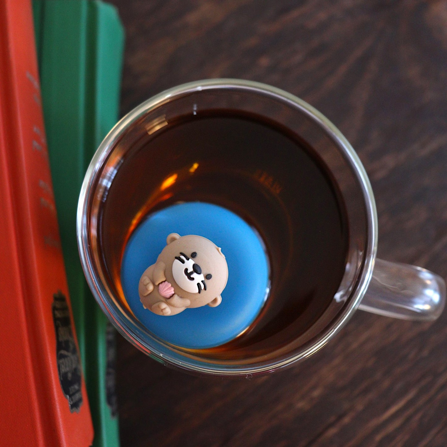 Looking down at a cute little otter floating on it's back holding a pink seashell tea infuser for loose leaf tea with a fine mesh stainless steel tea strainer that reads FLOAT2INFUSE on a blue base floating in a glass tea mug with books to the side