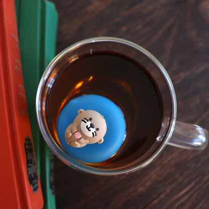 Looking down at a cute little otter floating on it's back holding a pink seashell tea infuser for loose leaf tea with a fine mesh stainless steel tea strainer that reads FLOAT2INFUSE on a blue base floating in a glass tea mug with books to the side