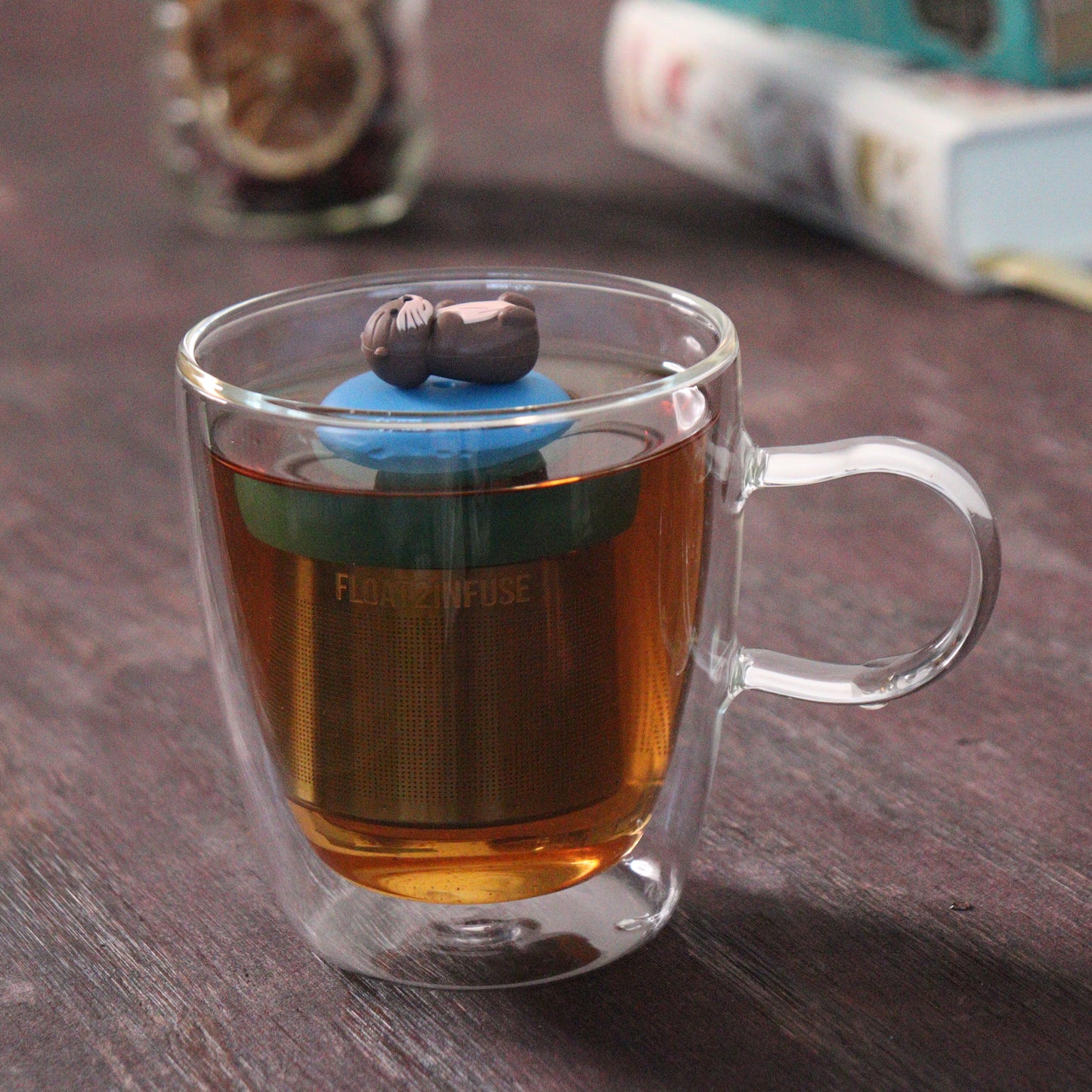 side view of an otter animal tea infuser for loose leaf tea  with a fine mesh stainless steel tea strainer that reads FLOAT2INFUSE on a blue base floating in a glass tea mug with books in the background