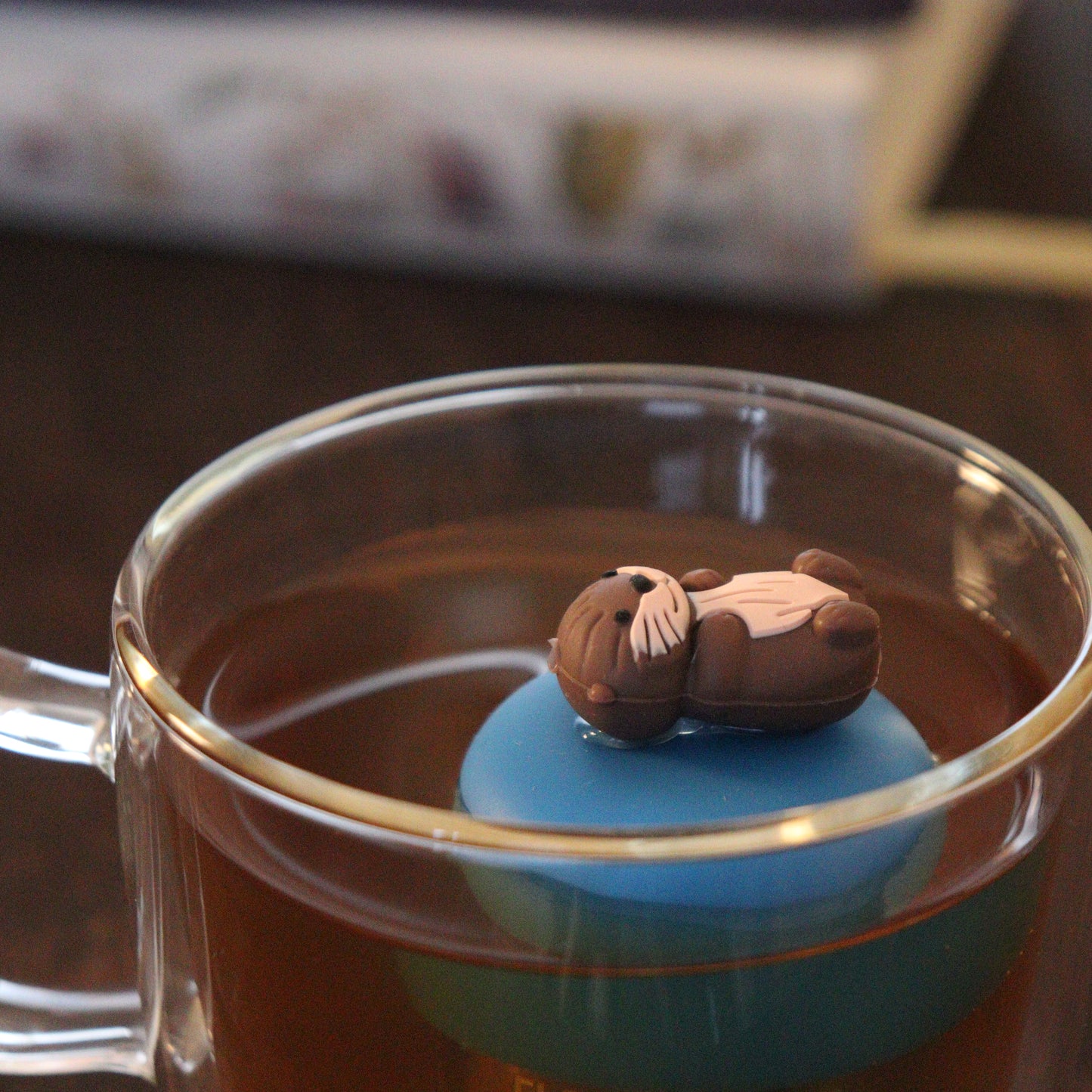 otter animal tea infuser for loose leaf tea with a fine mesh stainless steel tea strainer that reads FLOAT2INFUSE on a blue base floating in a glass tea mug  with books in the background