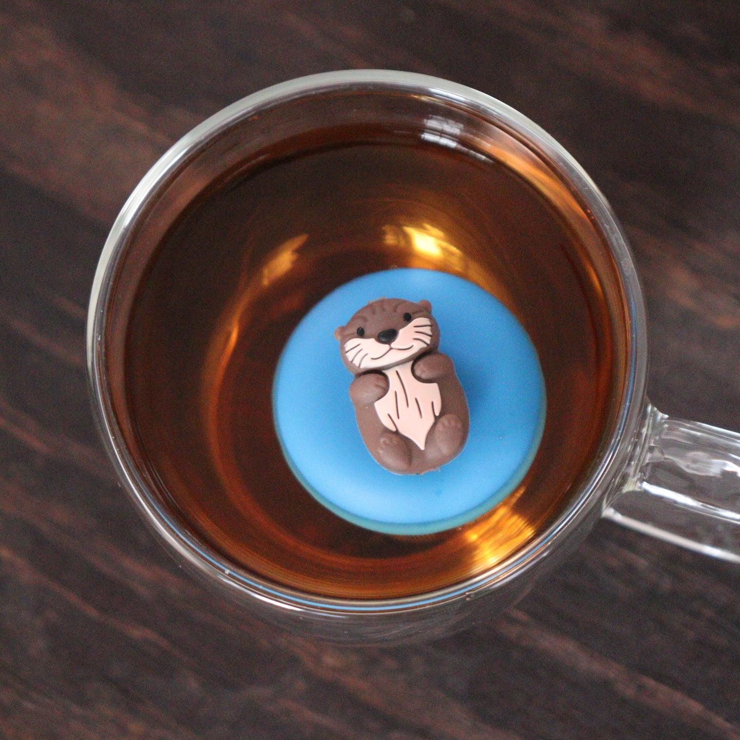 top view of an otter animal tea infuser for loose leaf tea with a fine mesh stainless steel tea strainer that reads FLOAT2INFUSE on a blue base floating in a glass tea cup 