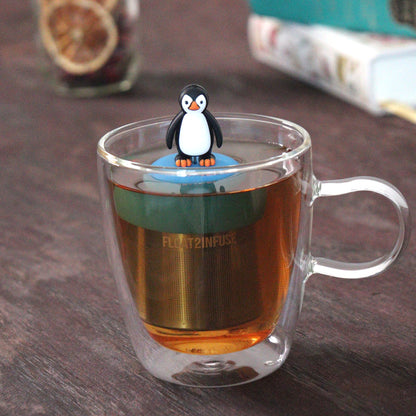 penguin tea infuser for loose leaf tea with a fine mesh stainless steel tea strainer that reads FLOAT2INFUSE on a blue base floating in a glass tea mug with loose leaf tea and books in the background