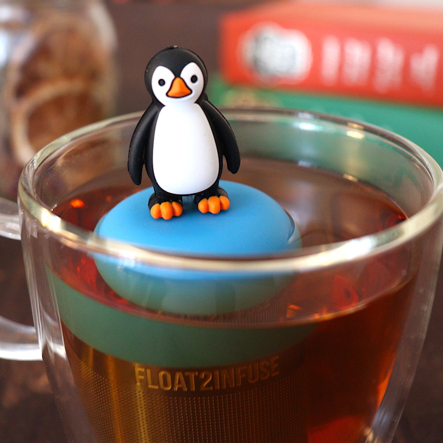 penguin tea infuser for loose leaf tea with a fine mesh stainless steel tea strainer that reads FLOAT2INFUSE on a blue base floating in a glass tea mug with books in the background
