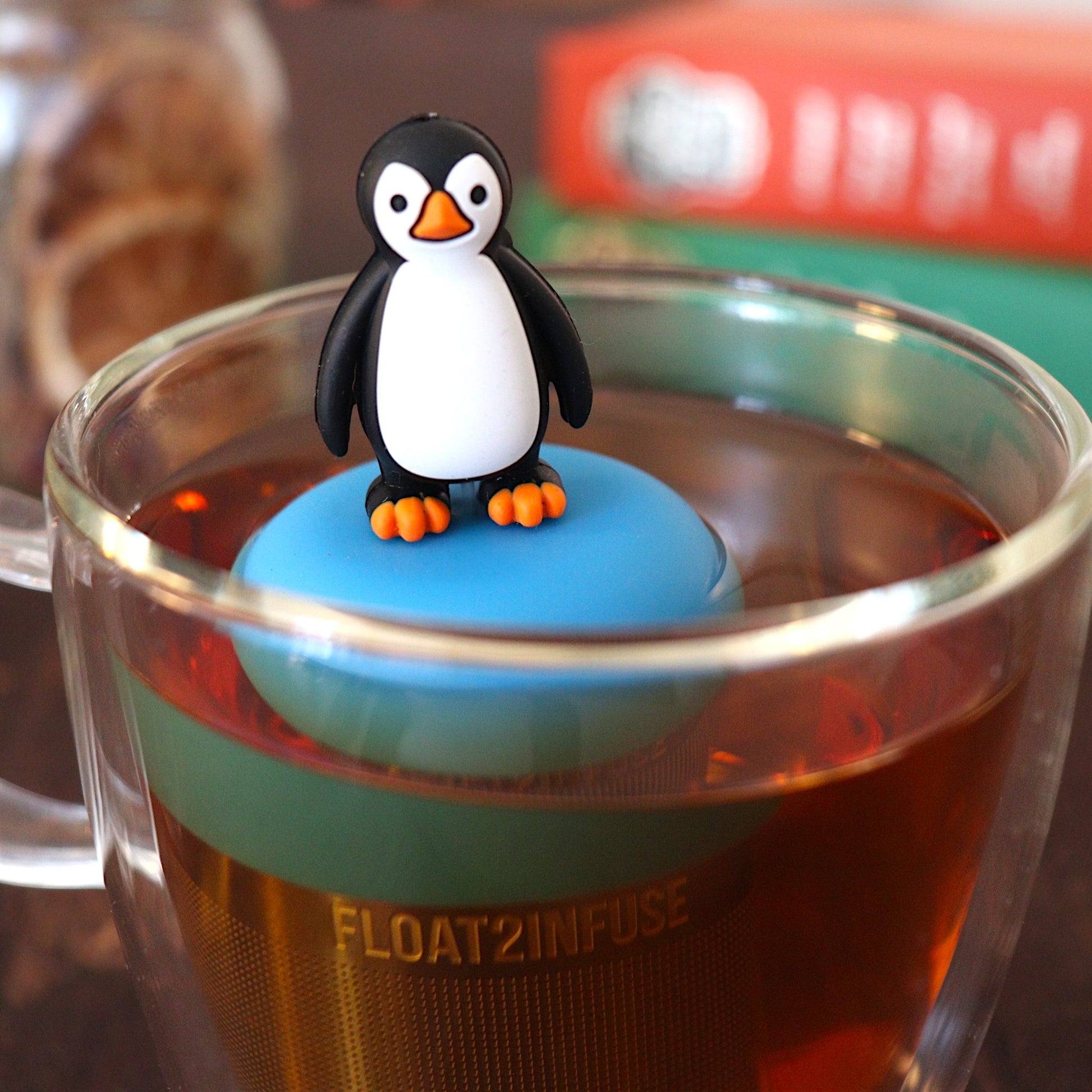 penguin tea infuser for loose leaf tea with a fine mesh stainless steel tea strainer that reads FLOAT2INFUSE on a blue base floating in a glass tea mug with books in the background